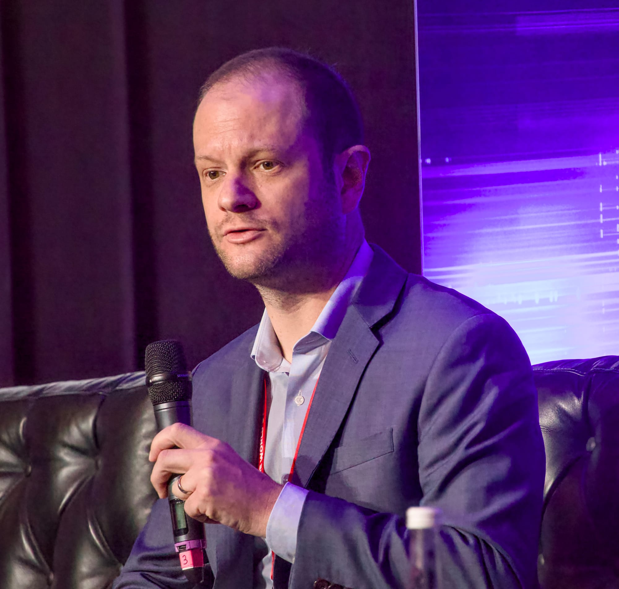 Matthew Monahan in a blue suit jacket and open-collared shirt sits on a dark leather sofa, holding a microphone in one hand. He wears a red lanyard and a wedding ring, speaking against a backdrop lit with purple light and abstract horizontal patterns, suggesting a conference or panel discussion.