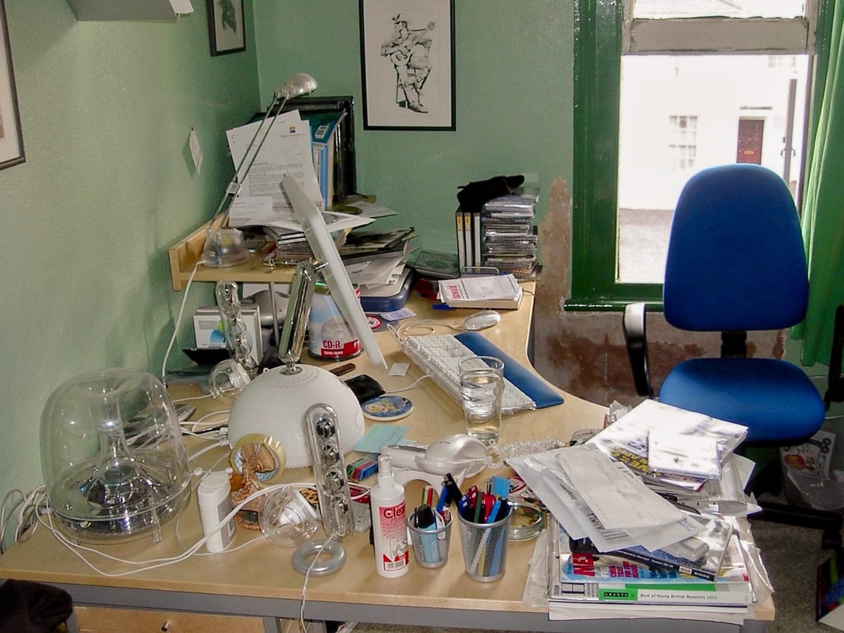 Cluttered home office desk with an old G4 iMac, scattered papers, CDs, pens, and a blue office chair by a window.