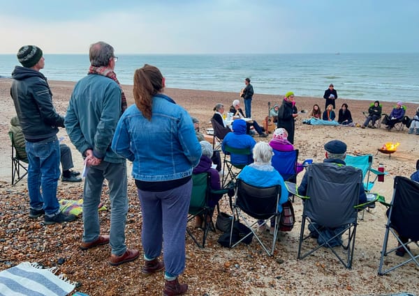Group of people gathered on a pebbled beach, many seated in camping chairs around a small fire, listening to speakers and musicians near the shoreline under a grey sky.