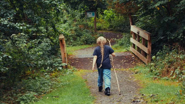 A child with a long braid walks on a forest path, holding a stick and approaching a small wooden bridge.