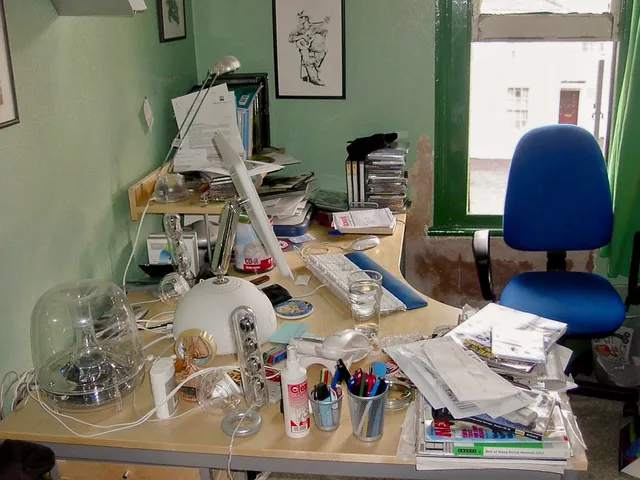Cluttered home office desk with an old G4 iMac, scattered papers, CDs, pens, and a blue office chair by a window.